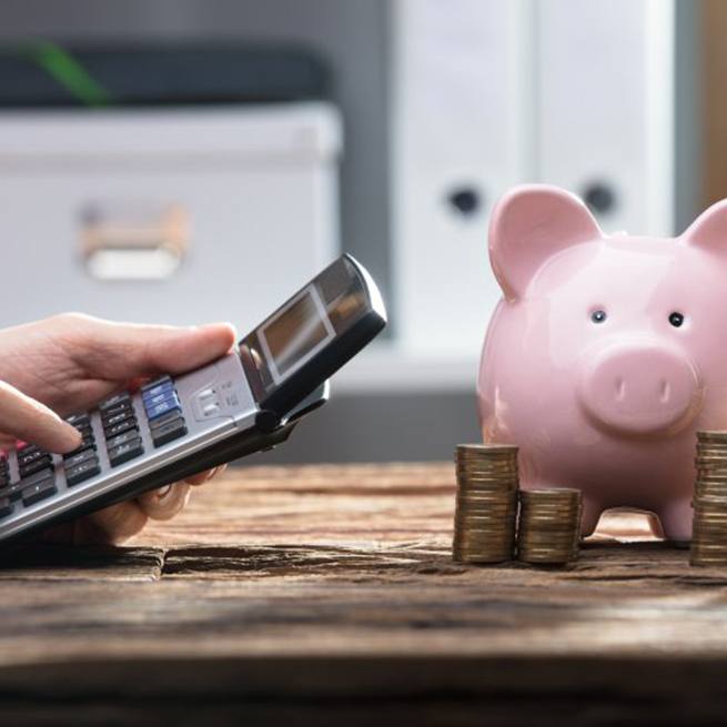 Individual using a calculator with piggy bank and coin stacks nearby